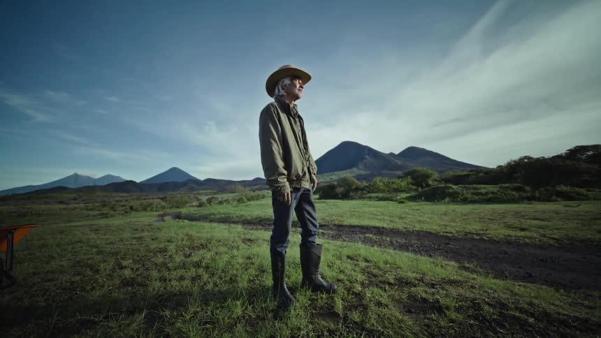 An old man in boots jeans and a jacket stands outdoors looking into the distance on a grassy plain