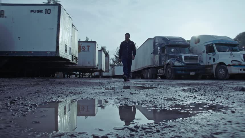 A man walking forward where there is a dirt floor with several water pits and there are some trucks parked behind