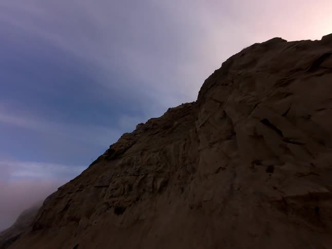 A steep sandy cliff bathed in the soft light of dusk with a hint of fog clinging to the base and a vast twilight sky above