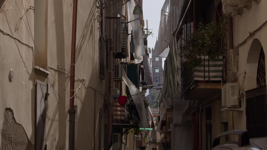 A narrow winding alleyway in a european city with tall buildings lining both sides and a glimpse of a distant church tower peeking through