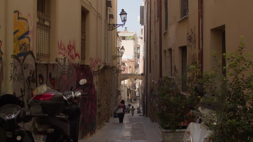 A narrow graffiticovered alleyway in a european city with a woman walking towards a bridge and a glimpse of a bustling street beyond