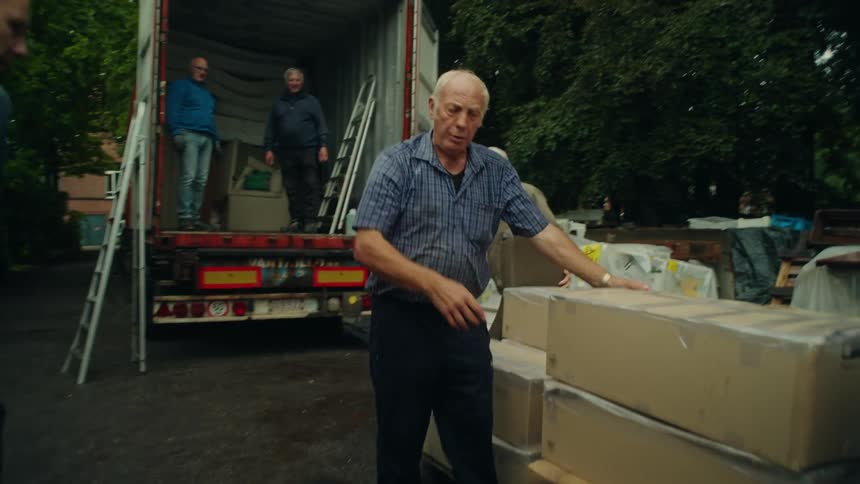 Some elderly men picking up some boxes that are next to a truck that has its back door open