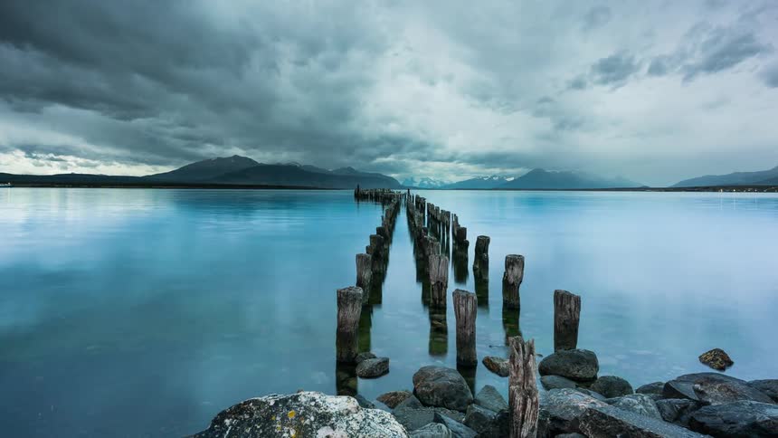 A row of weathered wooden pilings extends into a calm blue body of water with a dramatic cloudy sky and snowcapped mountains in the distance