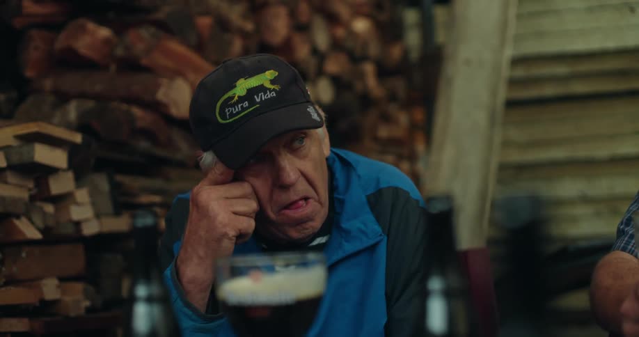 An elderly man wearing a black hat with a green animal drawn on it and he is looking forward and nodding at something while putting his hand on his face