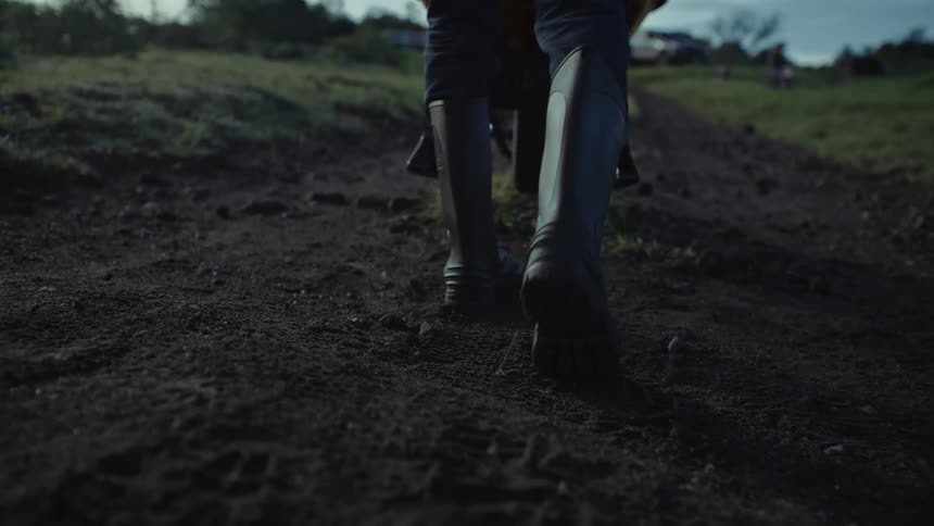 A person wearing boots carries a metal object down a dirt path in a rural area