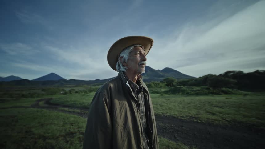 Elderly man in hat stands on hills with mountains in background