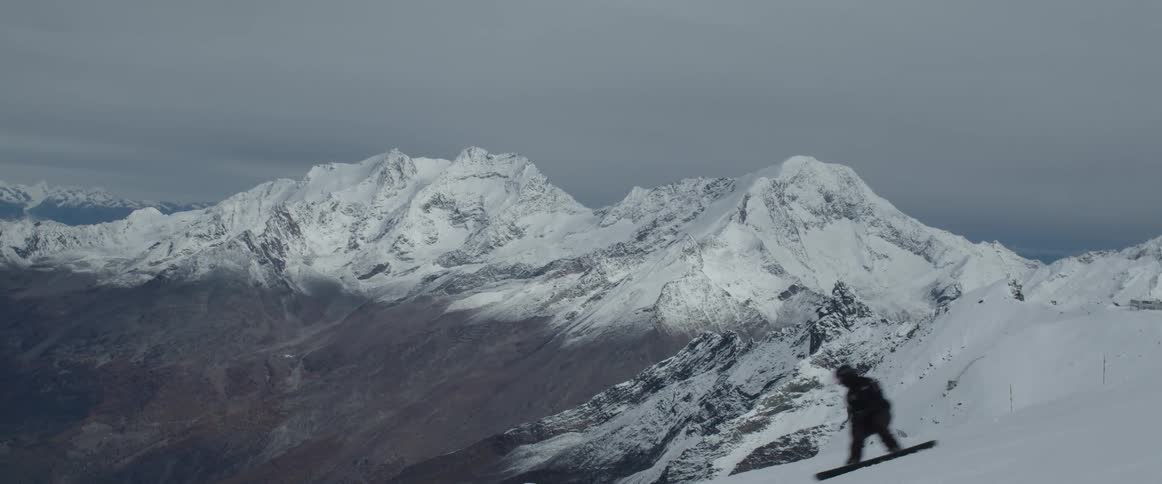 A man snowboards down a hill with a snowcapped mountain range in the background
