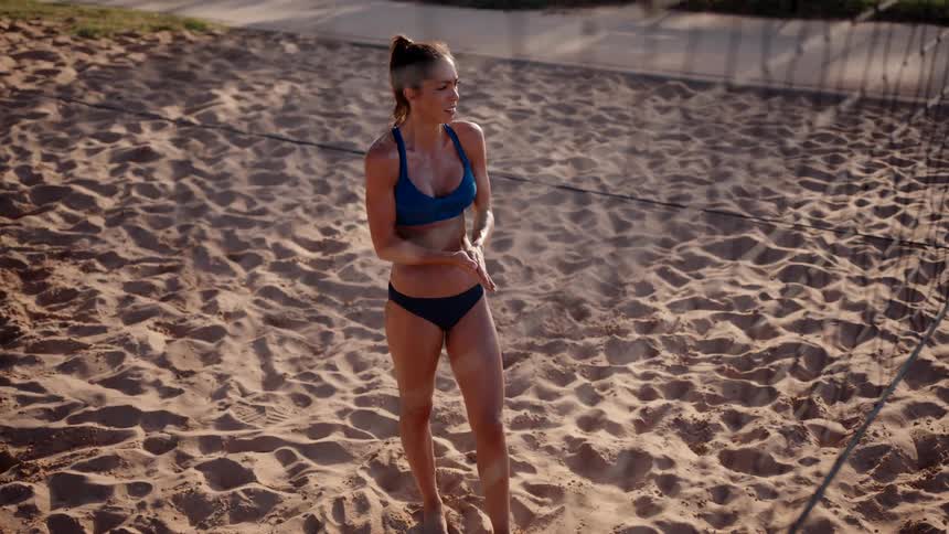 A young woman stands on the beach beside a volleyball net and rubs her hands together