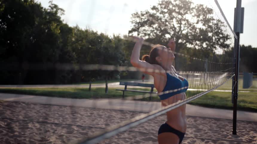 A woman jumps to hit a ball that is going over the net on a sand volleyball court
