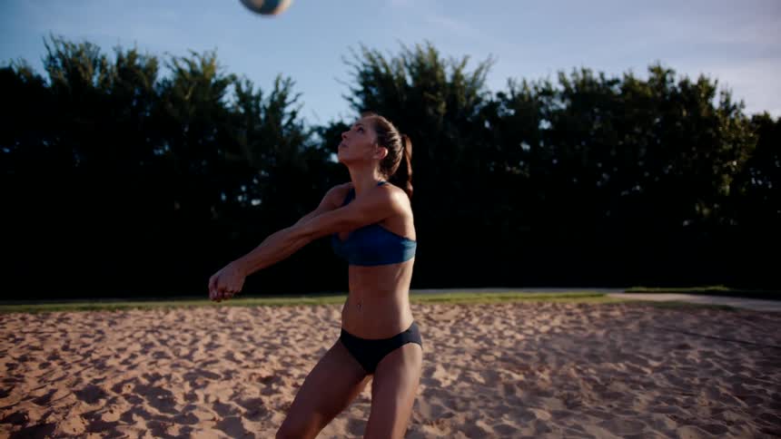 A woman in a bikini stands on the beach hitting a volleyball