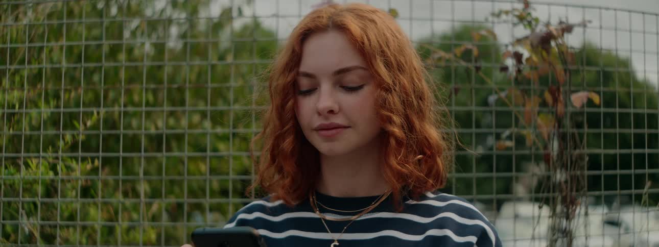 A redhead woman in a striped sweater browses her phone near a wire fence