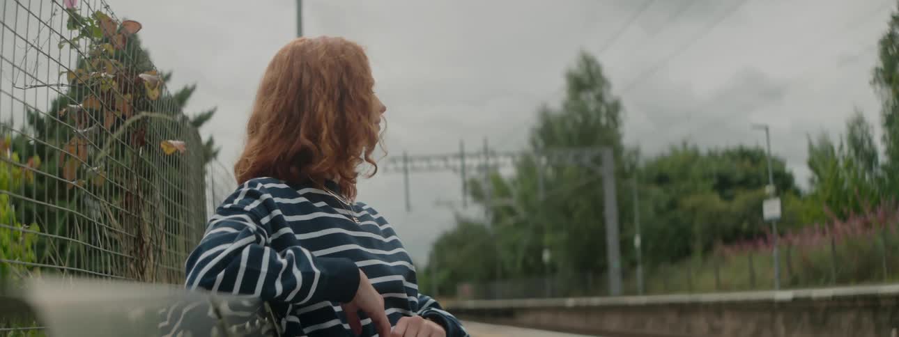Redheaded woman sitting near the railroad tracks turns her head to look down the line of tracks on an overcast day