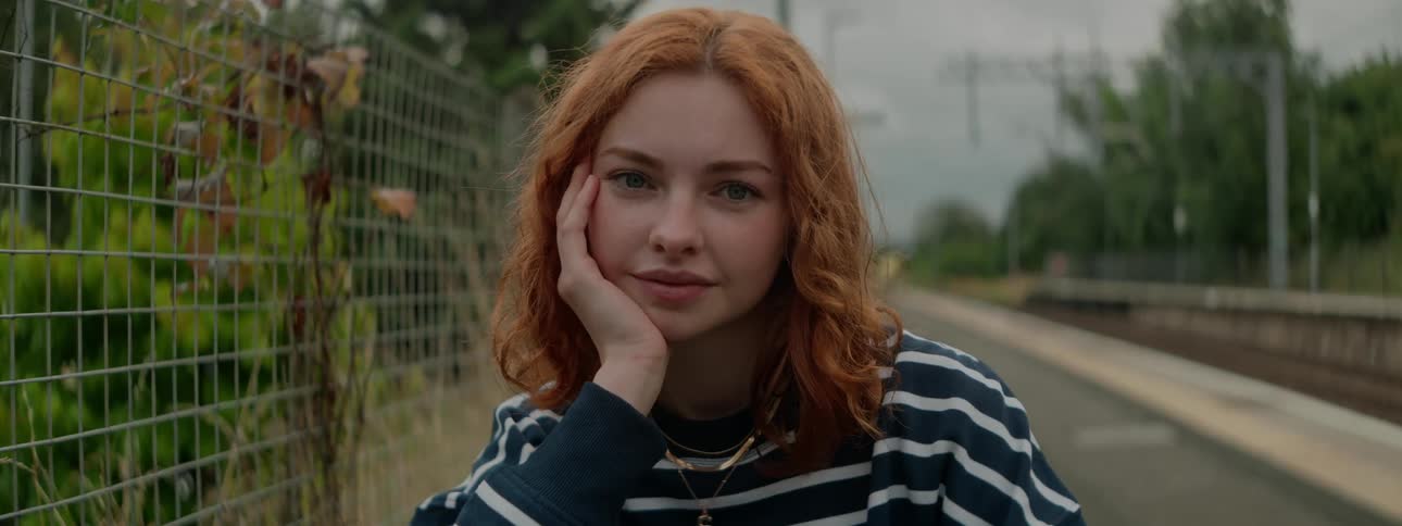 Redhead woman smiling on a road next to a fence with forest and foliage on the background outdoors during day time