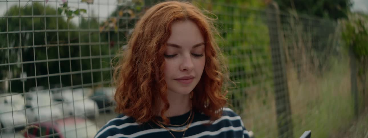 A red haired teenage girl stands next to a fence around a parking lot so she can read a book