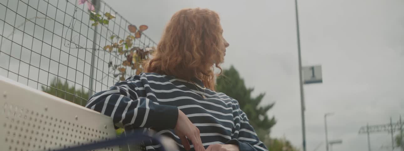 A young woman with short red hair and a striped shirt is sitting on an outside bench looking to her left and then looks to her other side