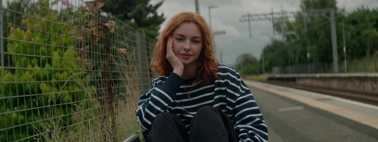 Redhead white woman sitting down on the floor next to a fence between a road and a forest outdoors during day time
