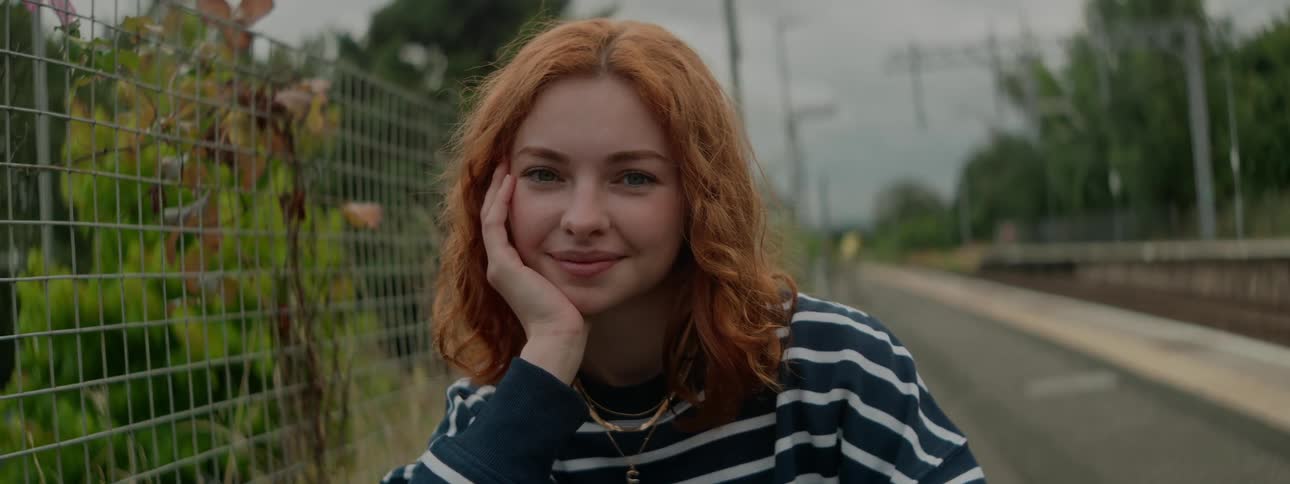 Smiling redhead poses with her head on her hand while sitting near the railroad tracks during the daytime