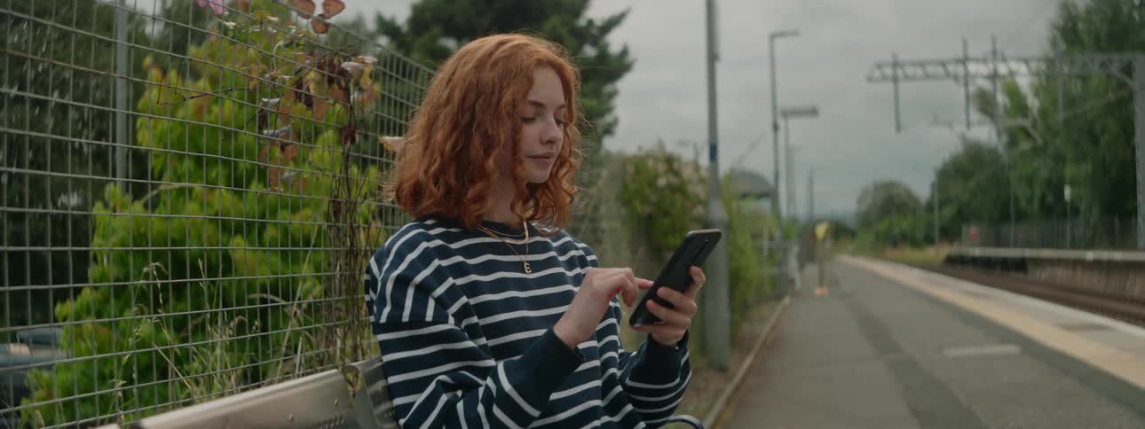 A redhead woman in a striped sweater browses her phone on the street