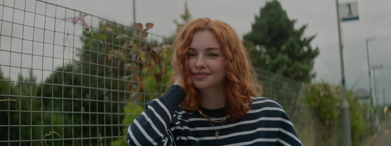 Redhead white woman standing next to a fence between a road and a forest smiling and touching her hair