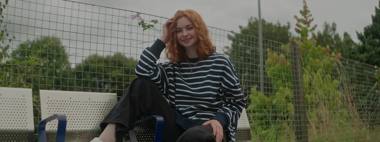 Redhead white woman sitting down on a bench next to a fence with a forest on the background outdoors during day time