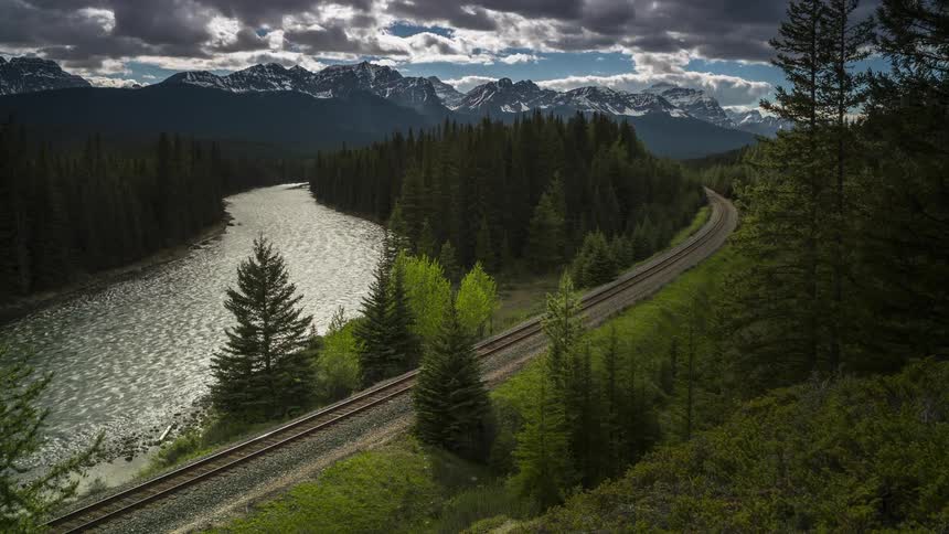 A train track winds through a lush forest alongside a river with snow capped mountains in the background