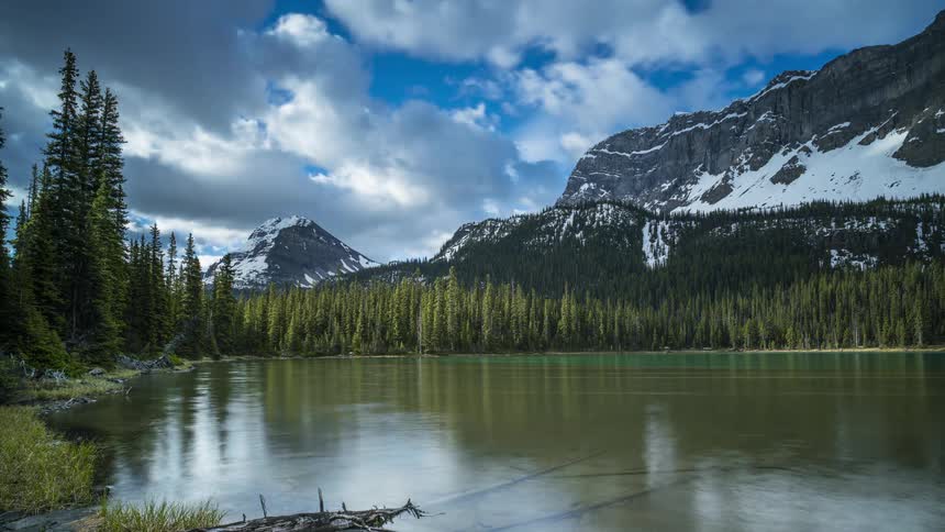 Passing clouds cast shadows onto a lake surrounded by forest and towered over by a mountain