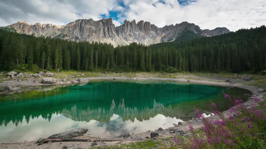 A lake surround by a thick forest and a mountain in the distance reflects the clouds floating overhead