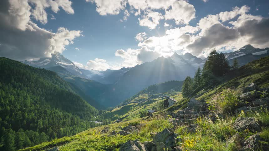 A wide shot of a valley in the swiss alps with a village nestled in the foothills and snow capped peaks in the distance bathed in sunlight