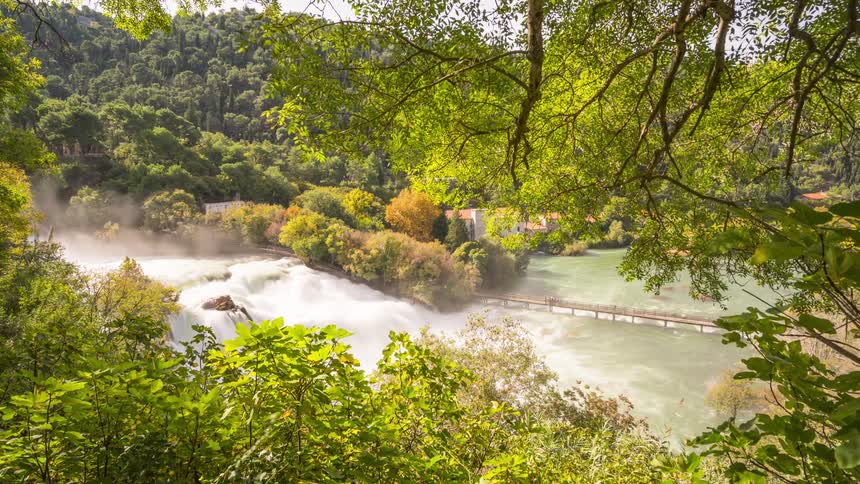 The skradinski buk waterfall in croatia with a wooden pedestrian bridge spanning the churning water framed by lush green foliage