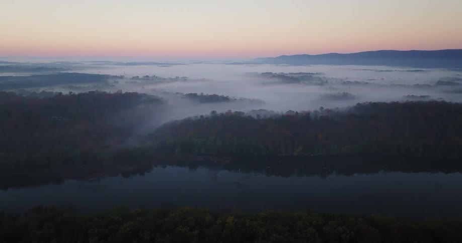 A wide aerial shot shows a river winding through a landscape of hills and forests covered in a thick layer of fog at dawn
