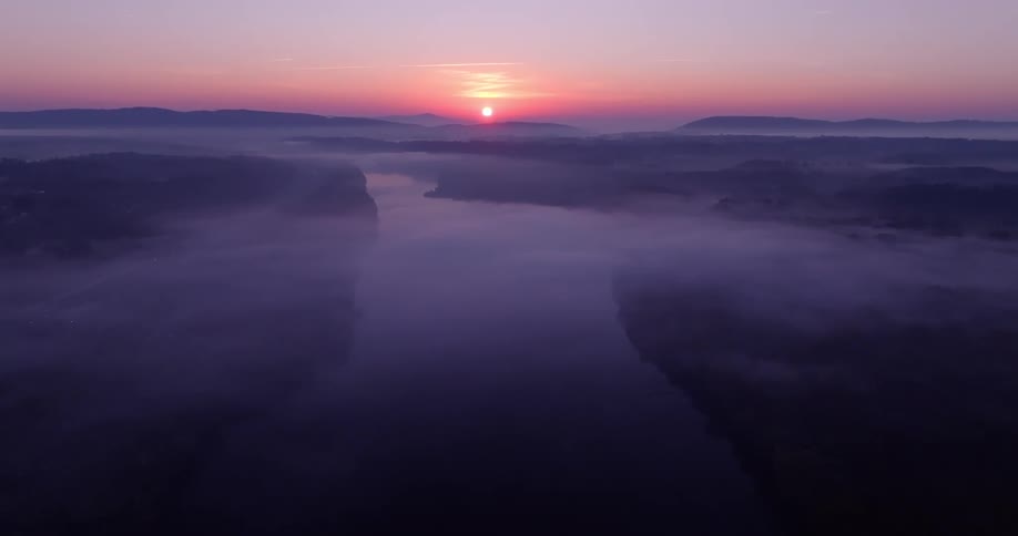 The river winds its way through the valley its path obscured by the swirling mist while the suns rays pierce through the clouds