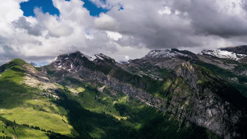 A mountainous landscape with rolling green hills and peaks some capped with snow under a dynamic sky filled with clouds