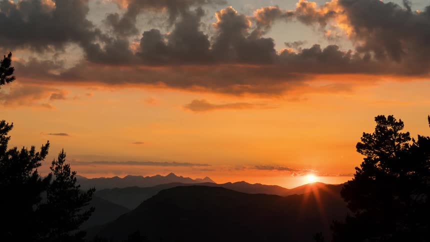 The sun sets behind mountain ranges as dark clouds roil in the sky above
