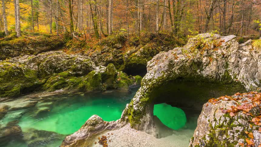 A crystal clear turquoise stream flows through a rocky autumnal forest with arches and openings in the rocks
