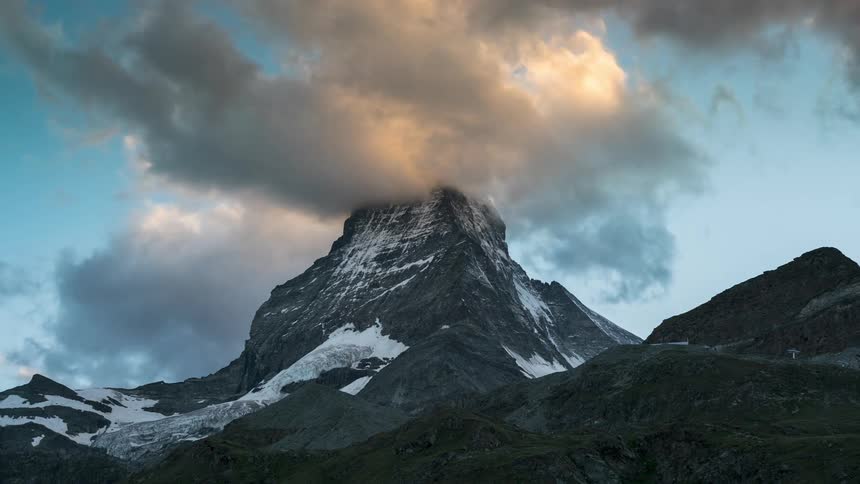 Dark clouds churn over a tall mountain peak partially covered in snow