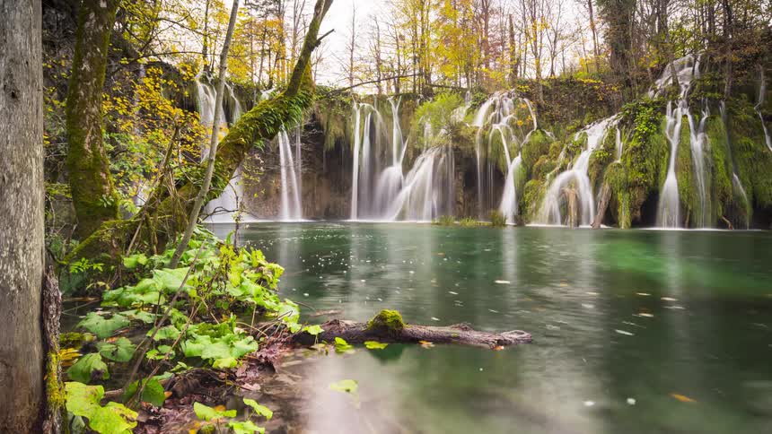 A serene autumn scene of cascading waterfalls into a placid pool of emerald green water surrounded by trees with vibrant fall colors