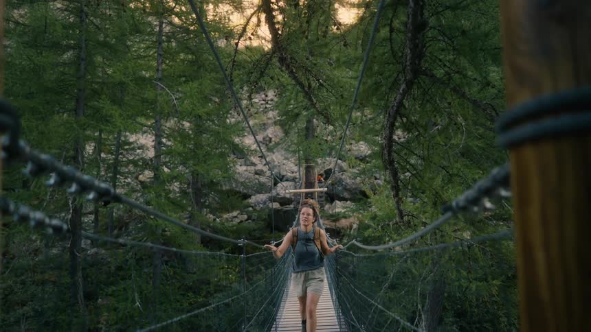 A woman wearing a backpack walks across a wooden suspension bridge in a lush forest