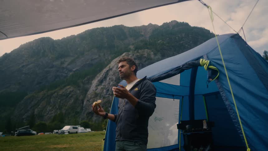 A man stands beside a blue tent eating and looking out at a mountain range
