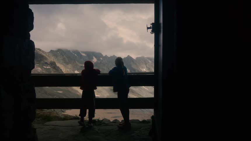 Two children look out over a mountainous landscape from a high peak on a cloudy day