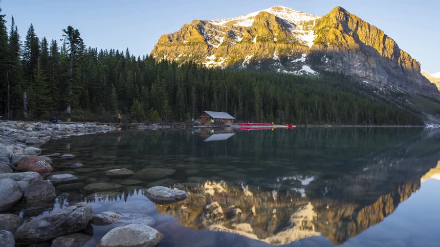 People approach and leave a small building that sits on a lakeshore near a forest and big mountain