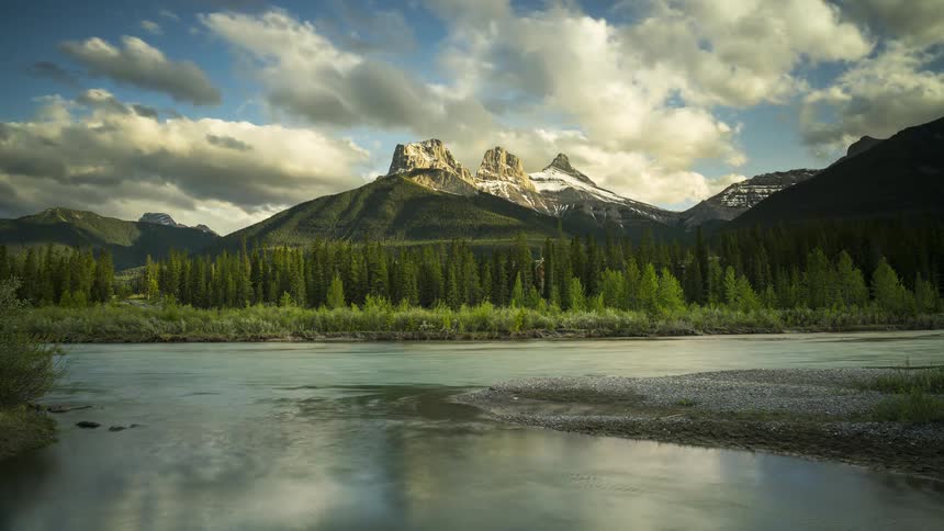 The three sisters mountain range rises over a calm river under rolling clouds