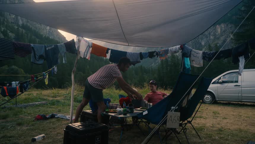 A group of three adults and two children are camping in a tent with clotheslines around it and one of the people is carrying a pot and placing it on a table