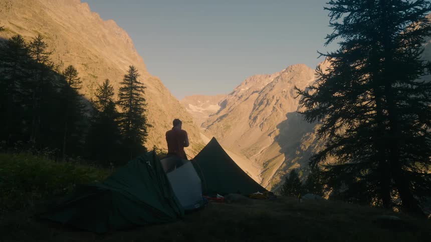 A lone hiker comes out of a tent amidst a mountain valley gazing at the majestic peaks bathed in the warm glow of the setting sun