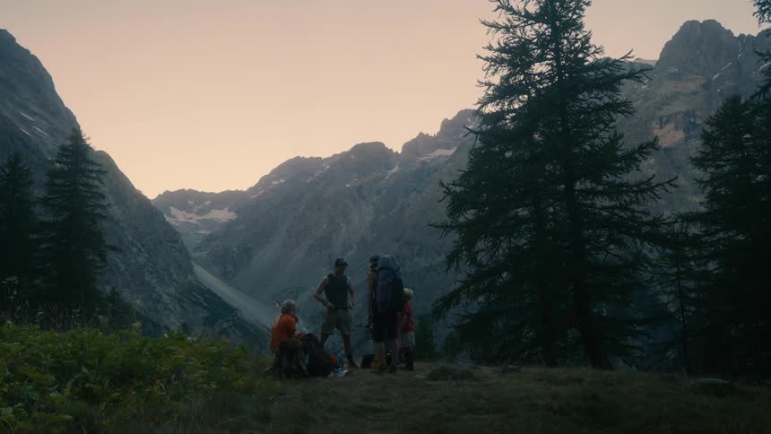 A group of hikers silhouetted against a backdrop of majestic mountains at dawn are gathered on a grassy hillside