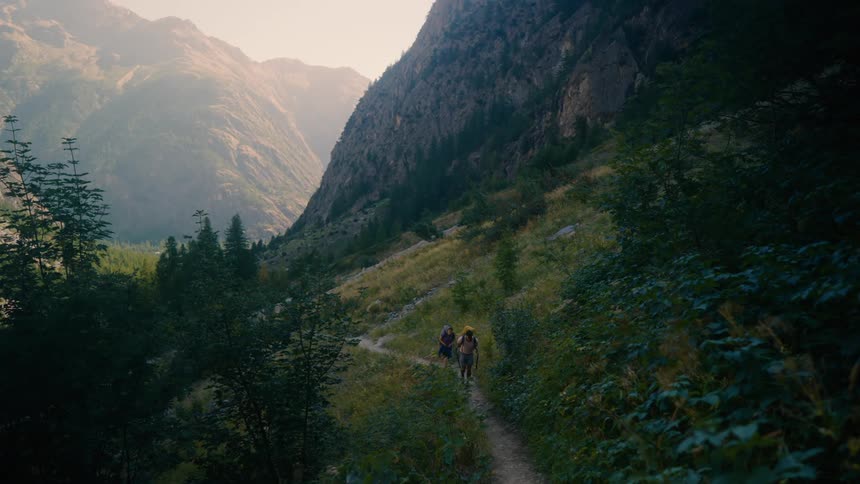 Two hikers with backpacks are seen walking along a mountain path surrounded by lush vegetation and towering mountains