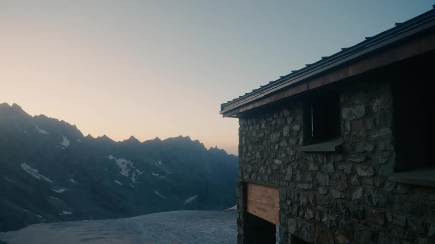 The rustic stone hut stands sentinel against a backdrop of snow capped mountains bathed in the soft glow of dawn