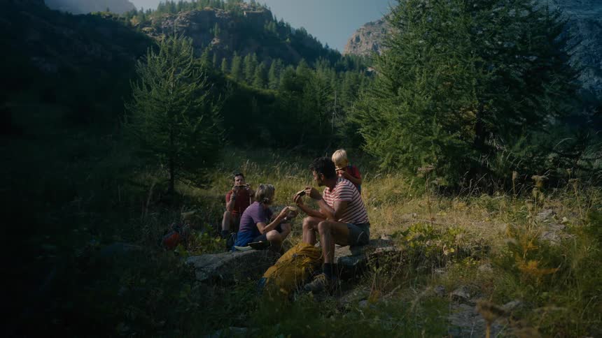 A group of people including a child are seated outdoors on rocks enjoying a meal in a mountain setting