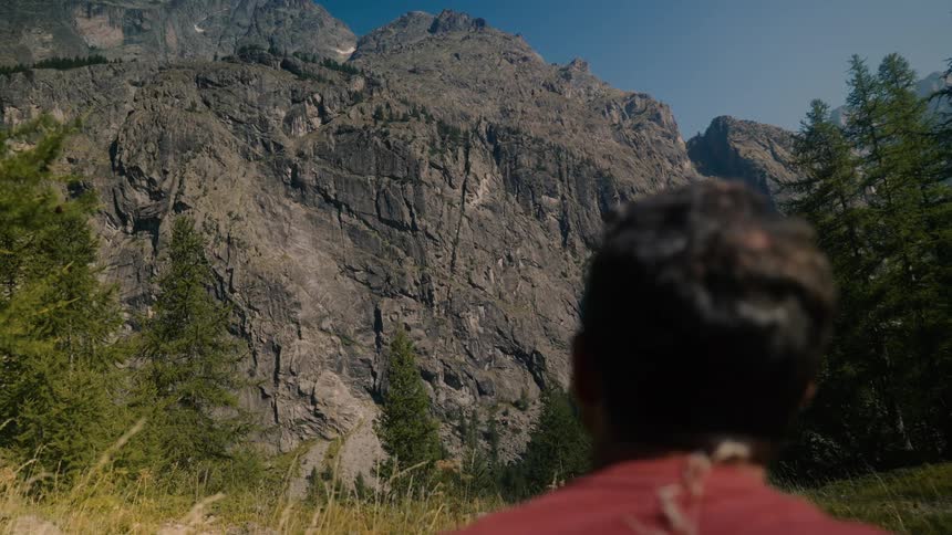 A man looks on as two hikers move down a path near a gigantic mountain range