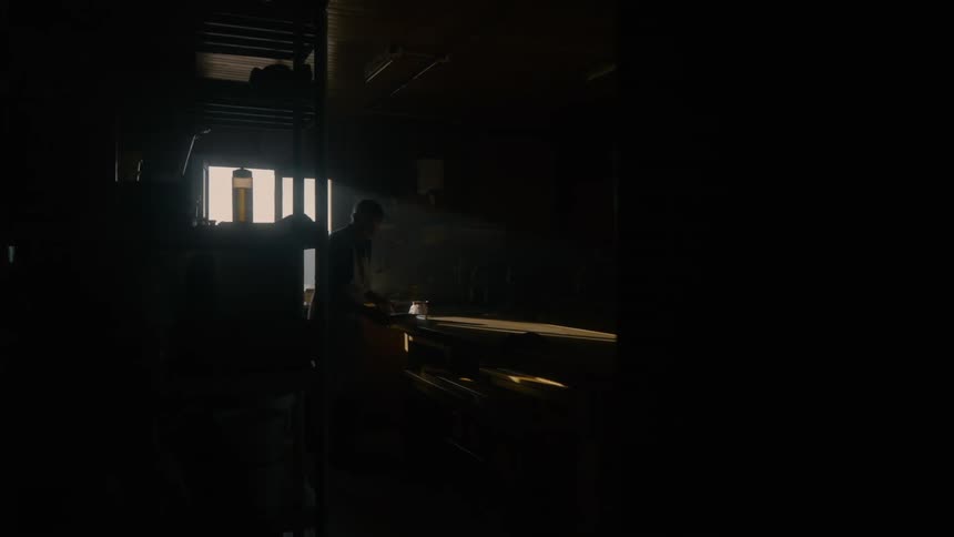A silhouetted carpenter works at a wooden workbench in a dark workshop illuminated by sunlight streaming through a window