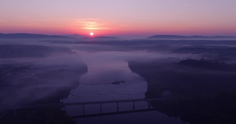 A bridge crosses a river in a rural area where the sun is setting and fog sits over land
